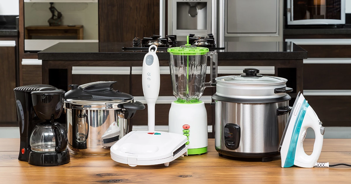 Imagem de alguns utencílios de cozinha posicionados um ao lado do outro, há uma cafeteira, uma panela de pressão, um mixer, um liquidificador, mais algumas panelas e um ferro de passar roupas, da esquerda para a direita.